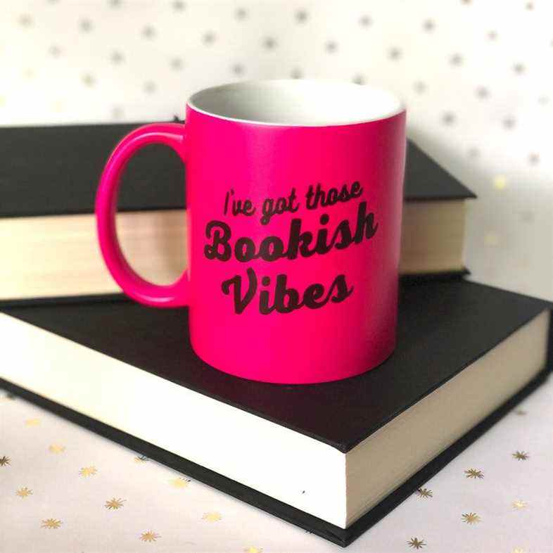 Mug rose vif avec les mots "J'ai ces vibrations livresques" dans une police noire rétro.  Il est posé sur un livre noir à couverture rigide. 