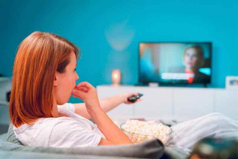 Young woman sitting on the bed sofa at home, lying with a popcorn bowl watching TV covered with blanket at her apartment alone enjoying movies or series holding remote control pointed