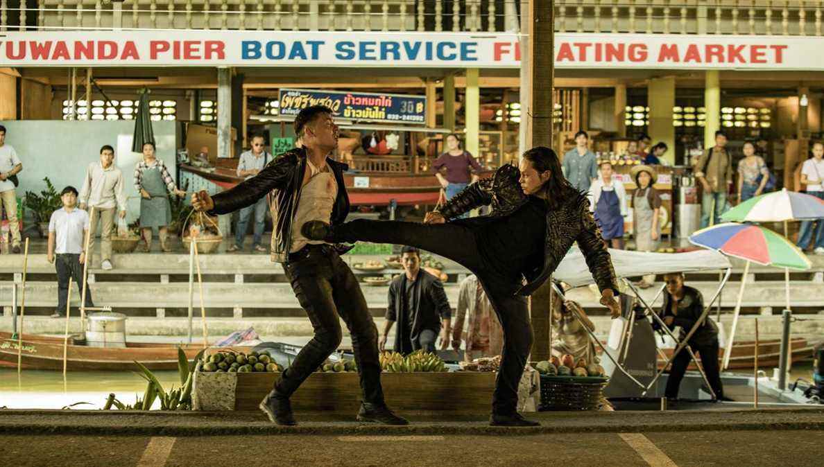 Lewis Tan, tout en noir avec une veste noire et dorée, donne un coup de pied total à un mec dans le ventre dans un marché en plein air dans Fistful of Vengeance