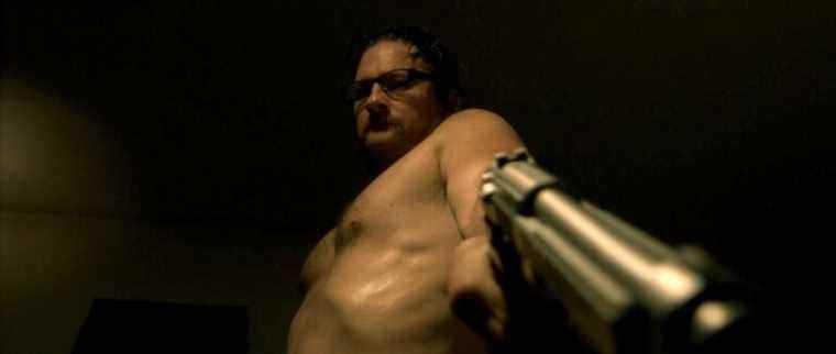 A shirtless Ray Belfi points a gun downward