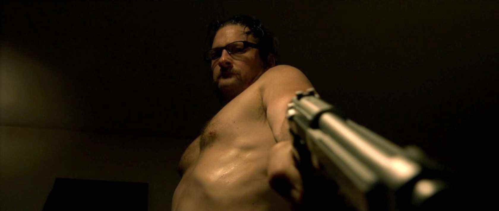 A shirtless Ray Belfi points a gun downward