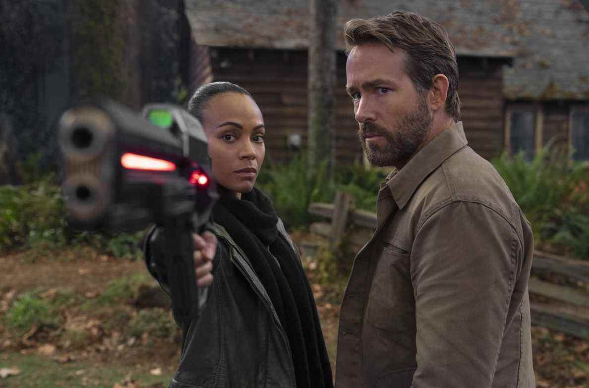 Zoe Saldana et Ryan Reynolds se tiennent côte à côte à l'extérieur dans The Adam Project, avec Zoe pointant un pistolet futuriste brillant vers la caméra