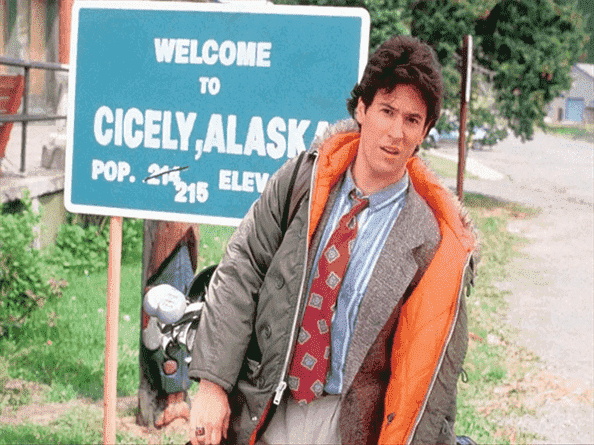 Rob Morrow dans Northern Exposure devant un panneau de bienvenue pour Cicely, Alaska.