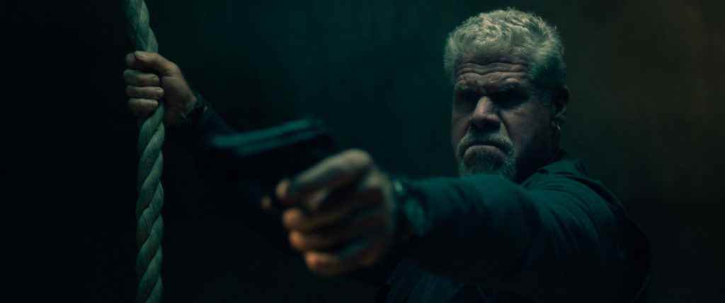 Ron Perlman pointant une arme à feu et tenant une corde dans There Are No Saints.