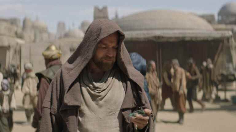 A middle-aged man with sandy hair and beard, wearing a brown hooded cloak and looking down at something in his hand as he stands in a desert city; still from "Obi-Wan Kenobi."