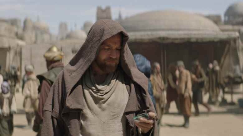 A middle-aged man with sandy hair and beard, wearing a brown hooded cloak and looking down at something in his hand as he stands in a desert city; still from "Obi-Wan Kenobi."