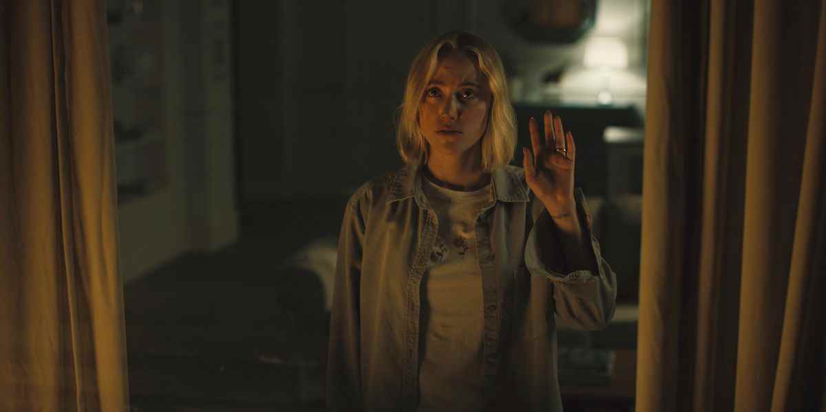 Maika Monroe se tient près d'une fenêtre, regardant vers l'extérieur, la main sur la vitre dans Watcher