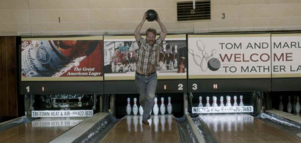 Wayne Stobierski (David Sullivan) courant dans une piste de bowling tenant une boule de bowling au-dessus de sa tête.