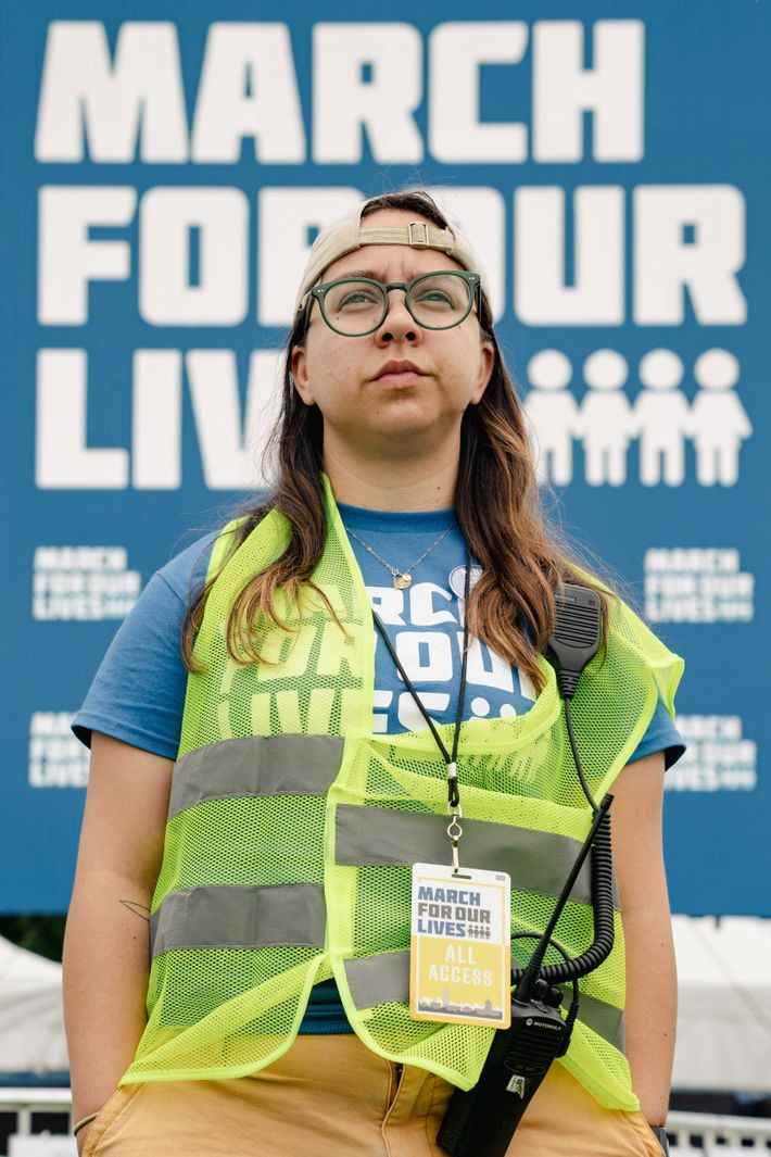 Une femme portant une casquette à l'envers, des lunettes vertes, une chemise March for our Lives et un gilet jaune fluo représente un portrait.