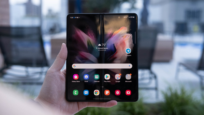 Une main tenant le Samsung Galaxy Z Fold 3 avec son écran ouvert et face à l'appareil photo.  Un pli court au milieu de la page d'accueil. 