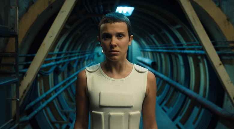 A teen girl with short buzzed hair wearing a white protective vest enters a mechanical hallway; still from "Stranger Things 4."