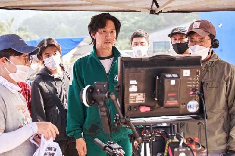 A man in a dark green tracksuit examines a television monitor, surrounded by others in masks; actor Lee Jung-jae behind the scenes of "Squid Game."