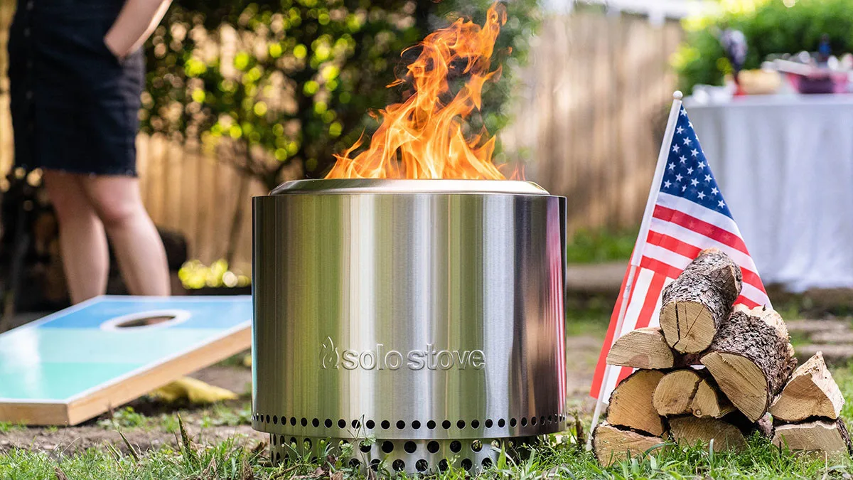 Un foyer Solo Stove assis sur l'herbe à côté de bois de chauffage et d'un drapeau américain lors d'une fête dans l'arrière-cour.