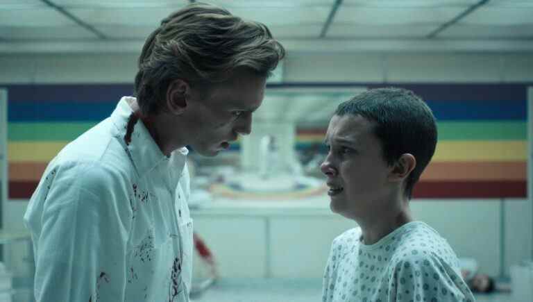 A blonde man in a white coat intimidates a teen girl with buzzed brown hair inside a playroom; still from "Stranger Things 4."