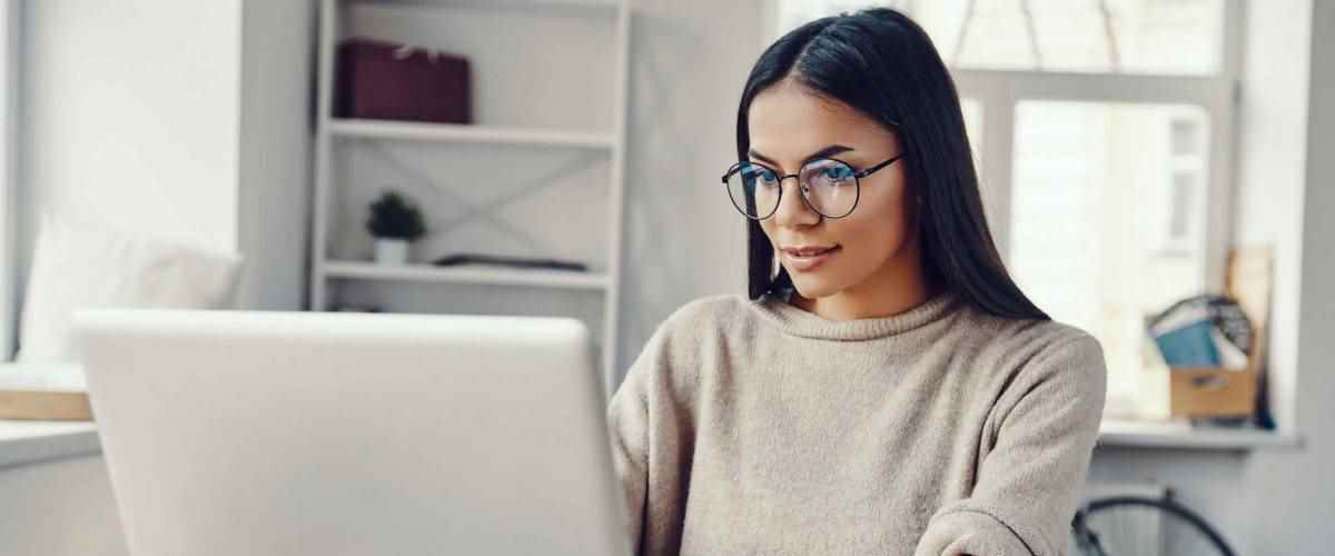 Belle jeune femme en vêtements décontractés utilisant un ordinateur portable et souriant tout en travaillant à l'intérieur