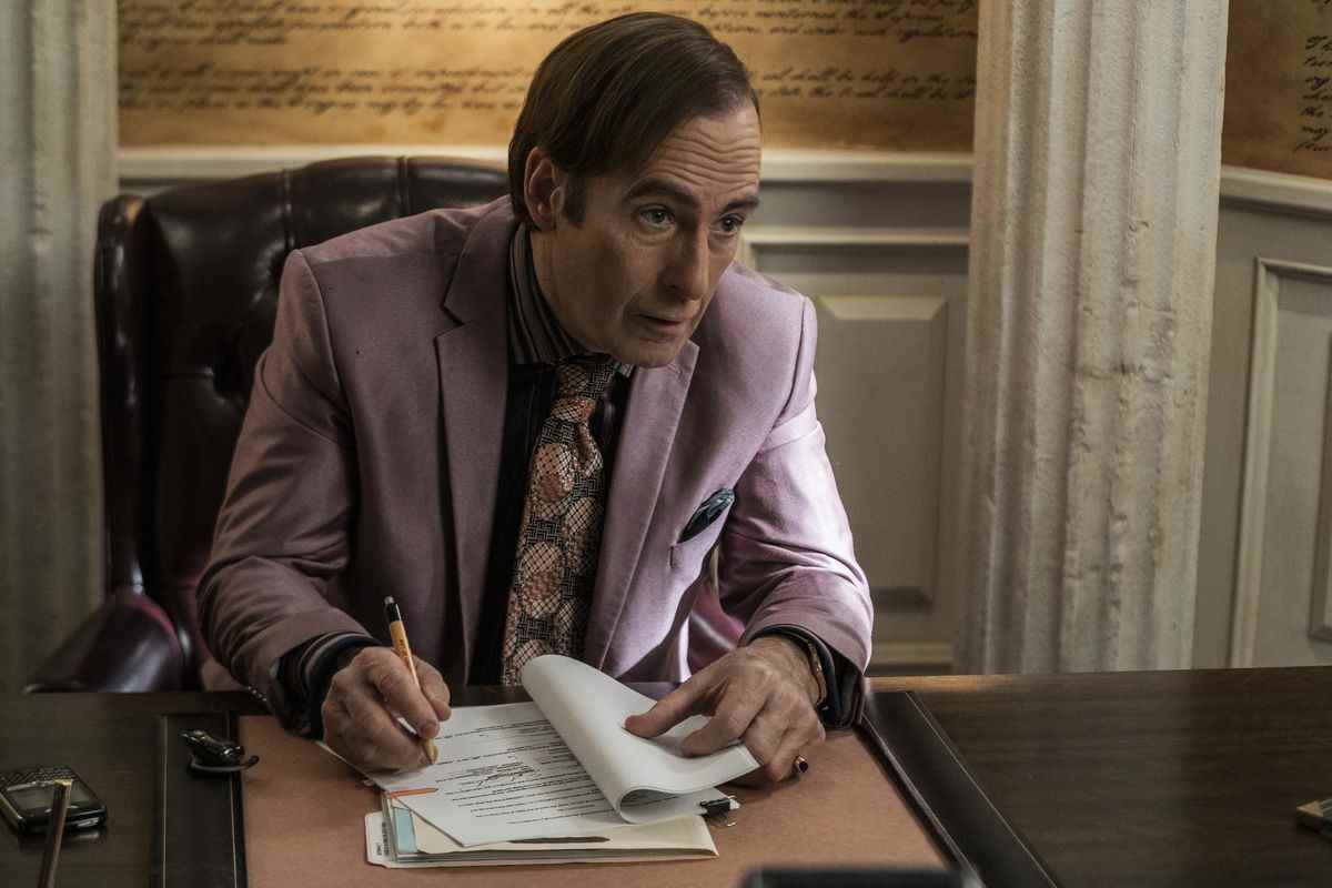 Saul Goodman assis à son bureau dans un costume violet regardant quelqu'un hors caméra.  La caméra est braquée sur lui en train de signer les papiers du divorce.