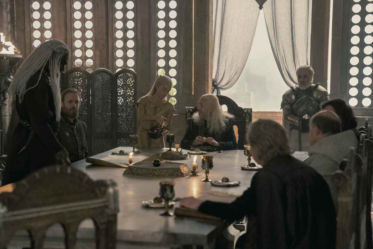 Viserys assis à sa table de conseil avec son conseil autour de la table et un garde derrière lui à la fenêtre.  Sa fille lui verse du vin et il la regarde