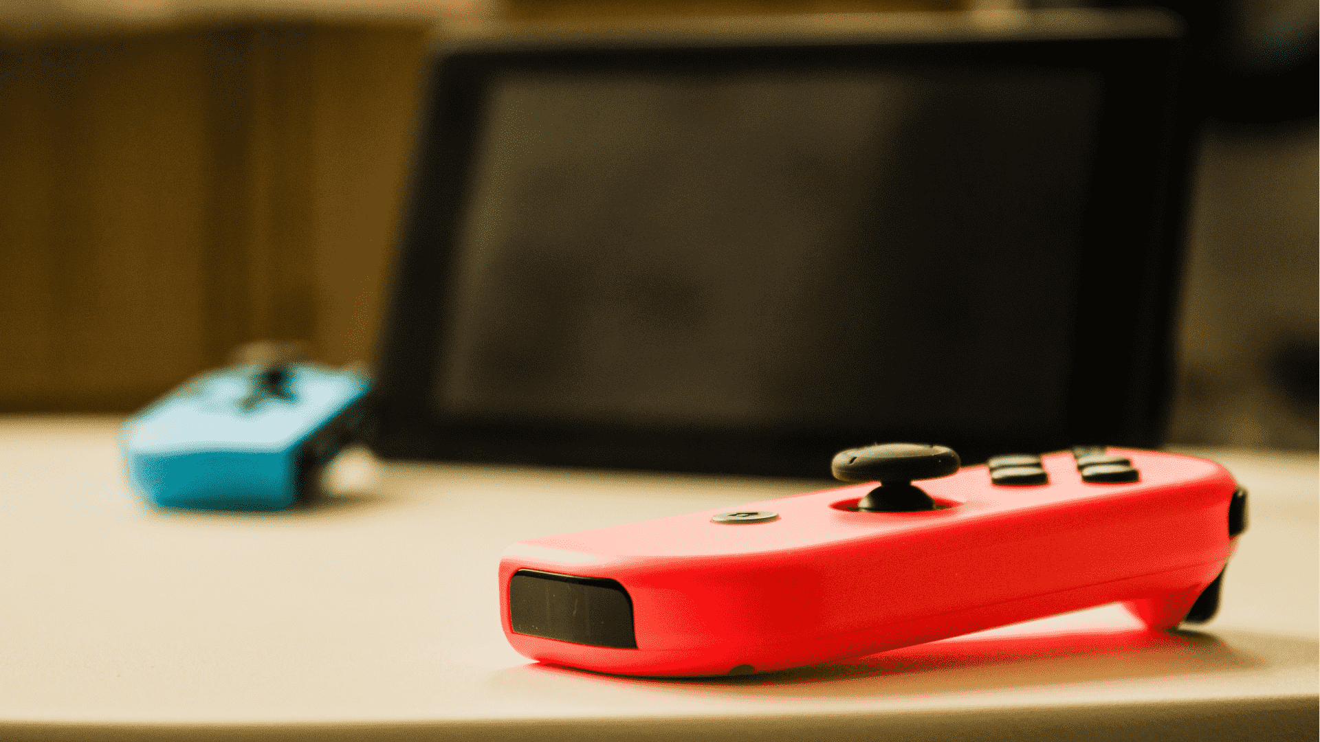 Un Joy-Con rouge fluo sur une table au premier plan, avec un écran Nintendo Switch et un Joy-Con bleu posé sur la table en arrière-plan, légèrement flou