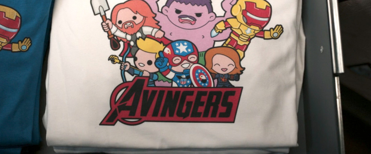 un t-shirt blanc avec des motifs chibi des personnages des vengeurs, mais tous légèrement décalés.  le hulk est violet, thor a les cheveux roux et en dessous il est écrit Avingers