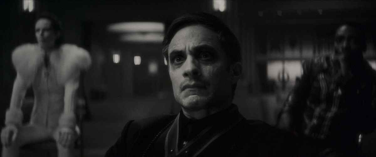 Gael García Bernal dans le rôle de Jack Russell assis les yeux écarquillés sur une chaise avec deux personnages d'horreur flous fabuleusement habillés derrière lui dans Werewolf by Night