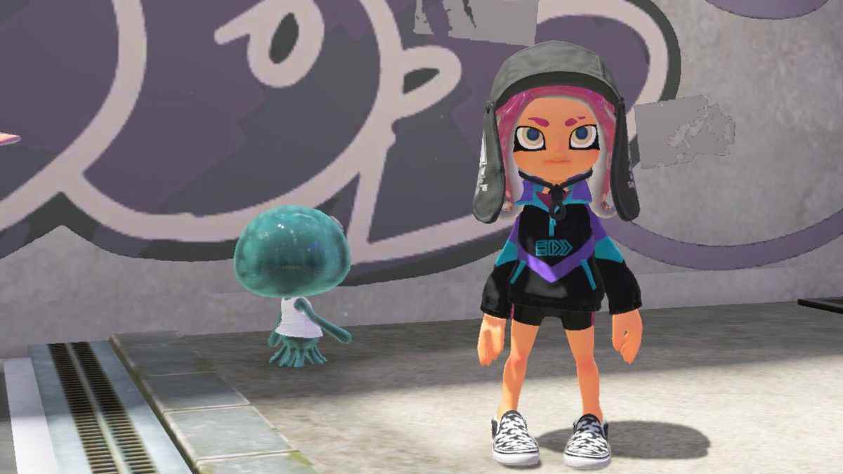 Un octoling aux cheveux roses se tenant devant un mur graffité dans Splatoon 3. L'octoling porte un chapeau noir avec des oreillettes, une veste en nylon de style années 80 contrastée en noir, aqua et violet, et pied de poule noir et blanc chaussures à enfiler.  Une méduse passe en arrière-plan.