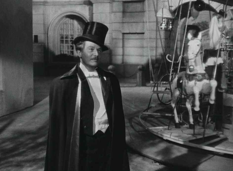 Anton Walbrook en tenue de maître de cérémonie à La Ronde, devant un carrousel.  Il porte un manteau noir fluide avec un chapeau haut de forme.