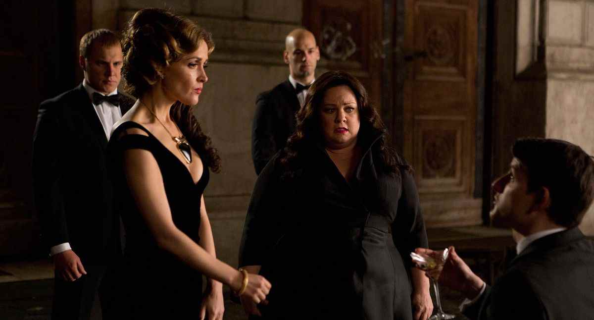 Melissa McCarthy et Rose Byrne portent des robes noires à l'extérieur dans Spy.  Ils se tiennent devant un homme agenouillé sur le sol et présentant quelque chose dans sa main.  Deux hommes à l'air sévère en smoking se tiennent derrière eux.