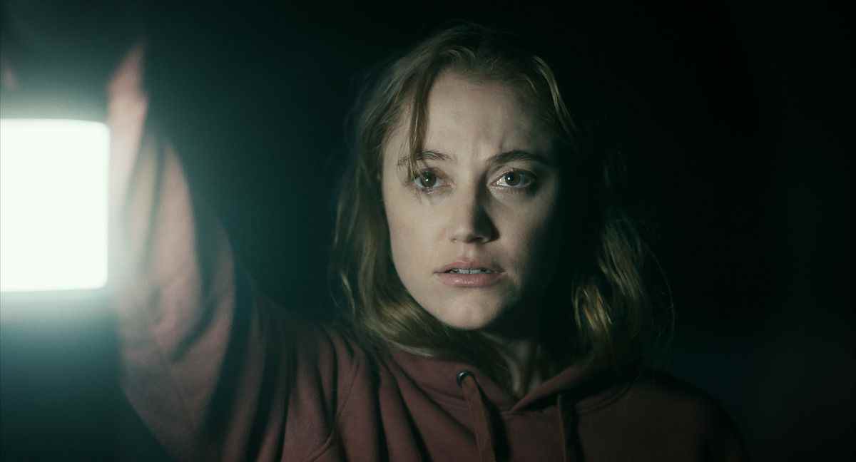 Une femme (Maika Monroe) portant un chandail rouge tient une lanterne dans une grotte sombre.