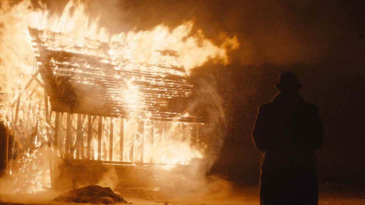 Un personnage vêtu d'un grand manteau d'hiver et d'une casquette melon se tient devant un bâtiment incendié au milieu de la nuit.