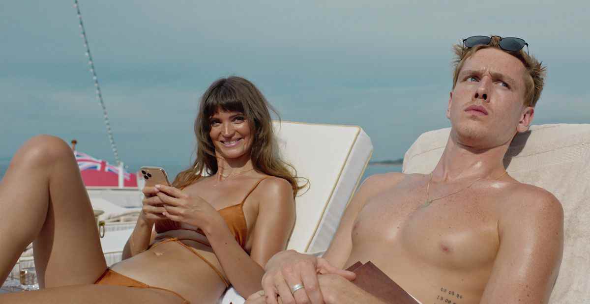 Les mannequins Yaya (Charlbi Dean) et Carl (Harris Dickinson) sont allongés en maillot de bain sur des chaises longues blanches sur le pont d'un yacht dans Triangle of Sadness, avec Yaya tenant son téléphone et souriant à quelque chose hors écran, et Carl ayant l'air perturbé