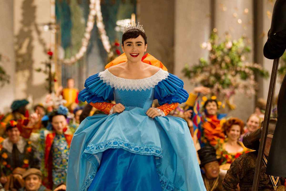 Blanche-Neige (Lily Collins) monte les escaliers de la salle du trône dans une somptueuse robe bleue avec une bordure blanche et un arc orange géant sur le dos alors que des personnes vêtues de costumes tout aussi colorés l'encouragent