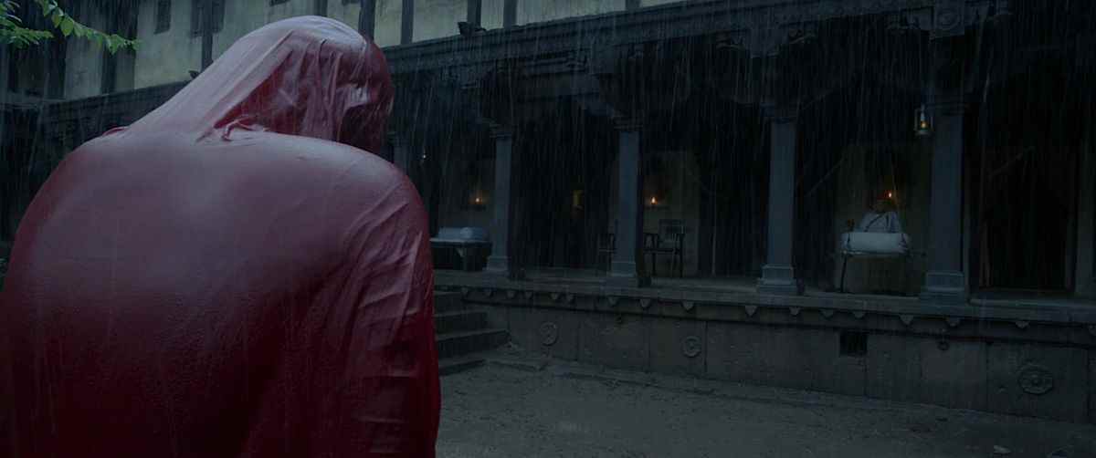 Une femme dans un sari rouge sang, mince et trempé se tient sous la pluie devant un porche couvert où un homme est assis et regarde dans le film d'horreur indien Tumbbad