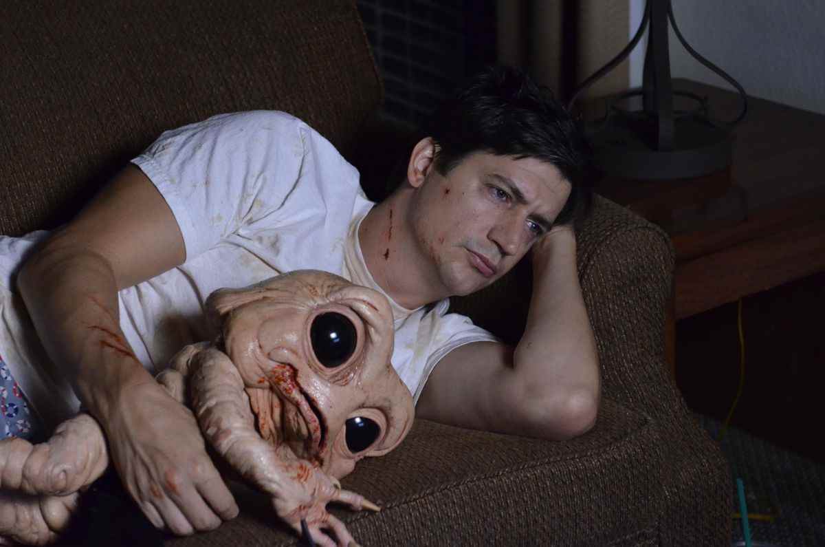 Duncan (Ken Marino) est allongé sur le canapé dans son t-shirt blanc tenant Milo, un démon des fesses qui ressemble vaguement au bébé de Dinosaures mais avec des yeux géants de Baby Yoda