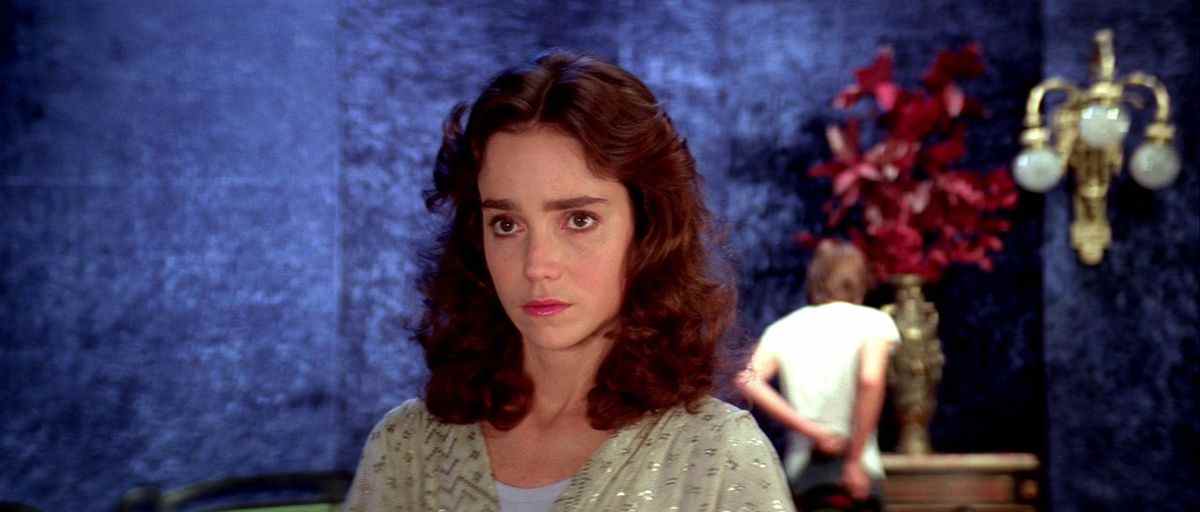 Jessica Harper dans Suspiria, avec un mur bleu, des fleurs rouges et un homme en chemise blanche.