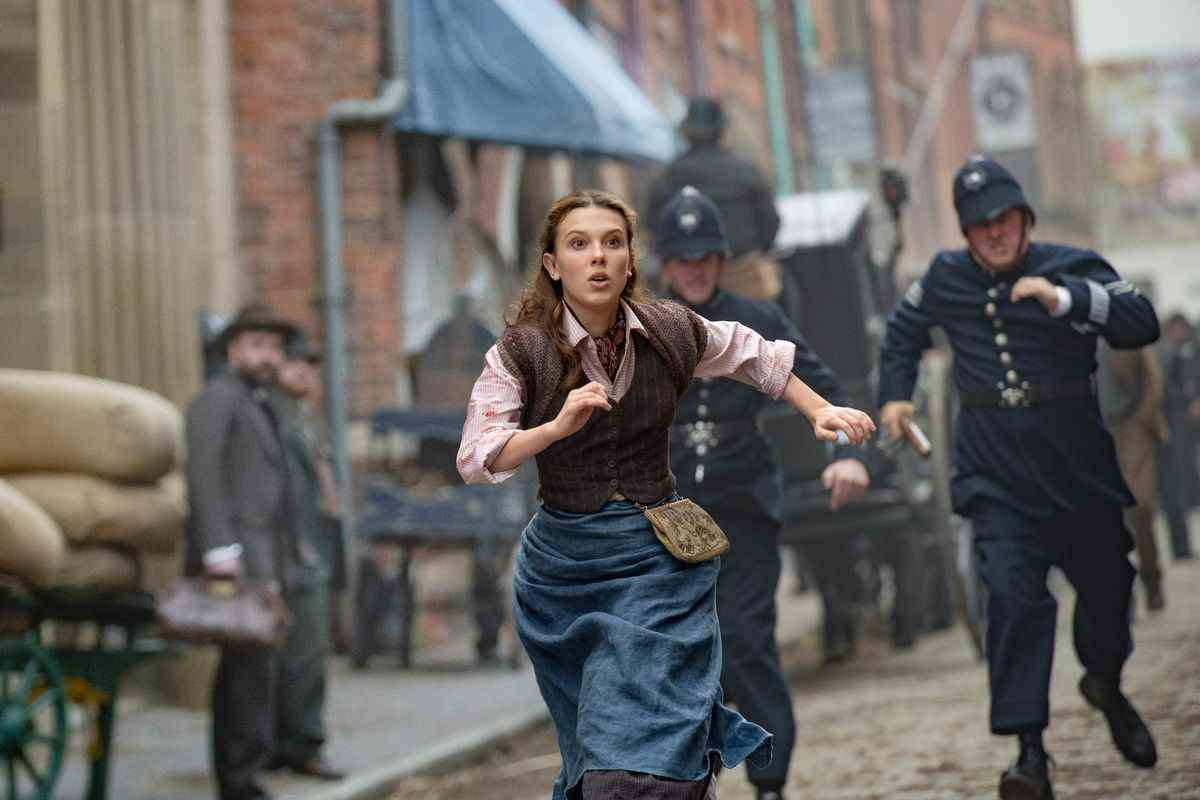 millie bobby brown dans le rôle d'enola holmes.  elle court dans les rues de Londres, deux policiers à ses trousses