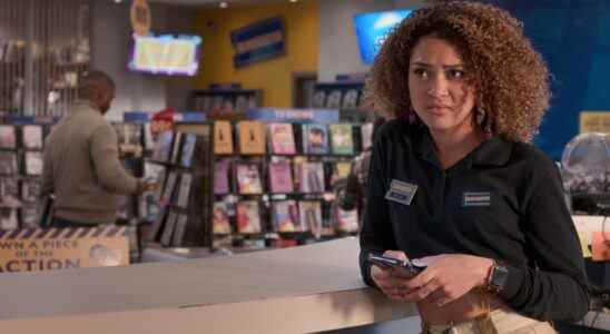 A teenage girl with curly brown hair in a navy blue Blockbuster Video shirt and khakis, standing holding her phone and looking disinterested in a Blockbuster video store; still from "Blockbuster."