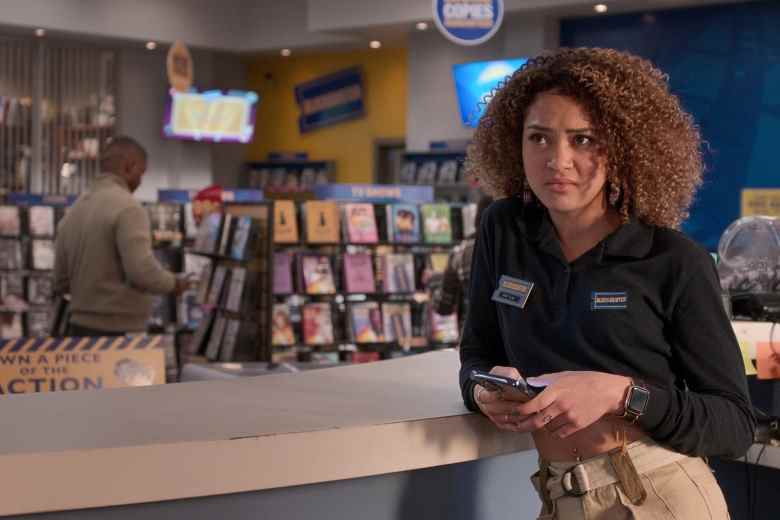 A teenage girl with curly brown hair in a navy blue Blockbuster Video shirt and khakis, standing holding her phone and looking disinterested in a Blockbuster video store; still from "Blockbuster."