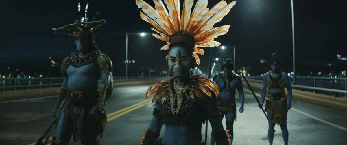 Un groupe de guerriers Talokan à la peau bleue se tient au milieu d'un pont la nuit, l'un d'eux portant un casque à plumes orange élaboré, dans Black Panther: Wakanda Forever.