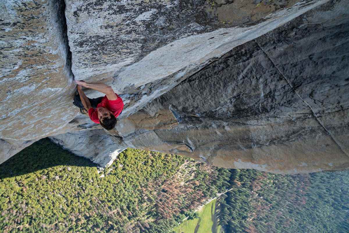 Un homme en t-shirt rouge et short bleu s'accroche à une crevasse face à une montagne escarpée avec une forêt d'arbres visible en contrebas.