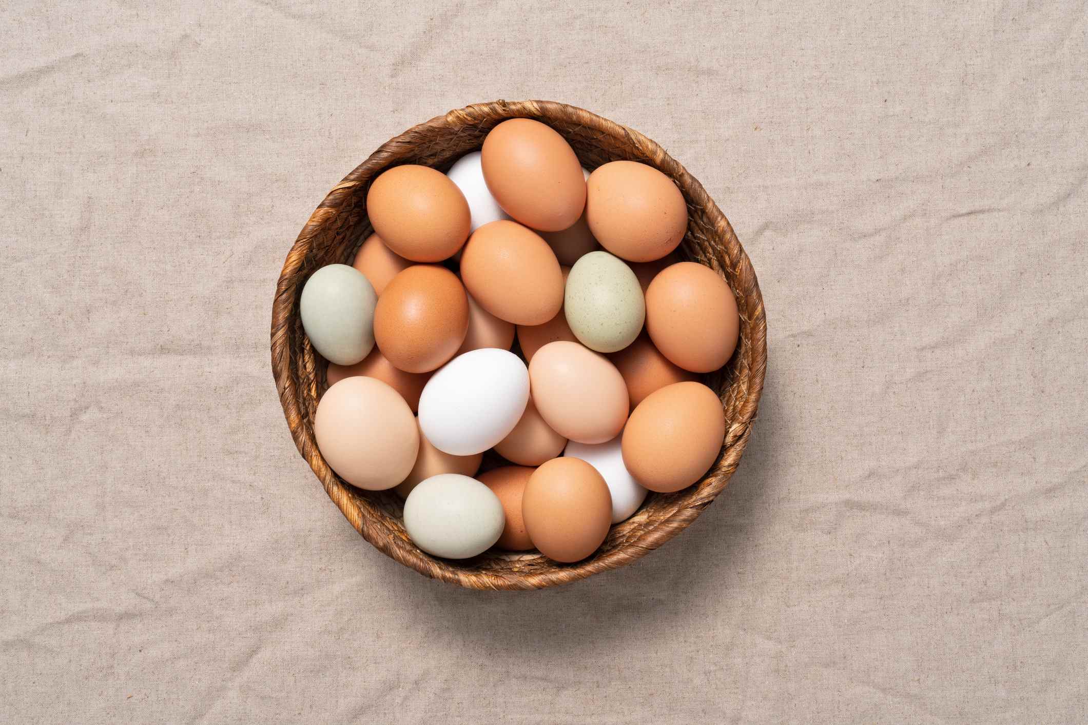 Oeufs de poule multicolores dans un panier sur nappe en lin de couleur beige.