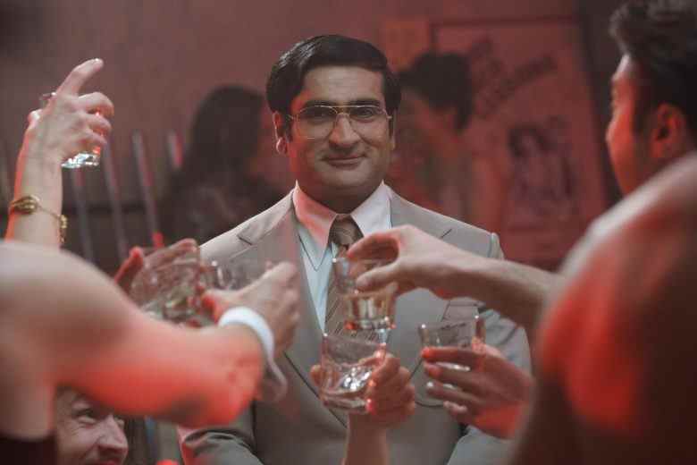 A man in a tan 1970s suit and large wire-rimmed glasses raising a glass along with others (only their hands in frame); still from "Welcome to Chippendales"