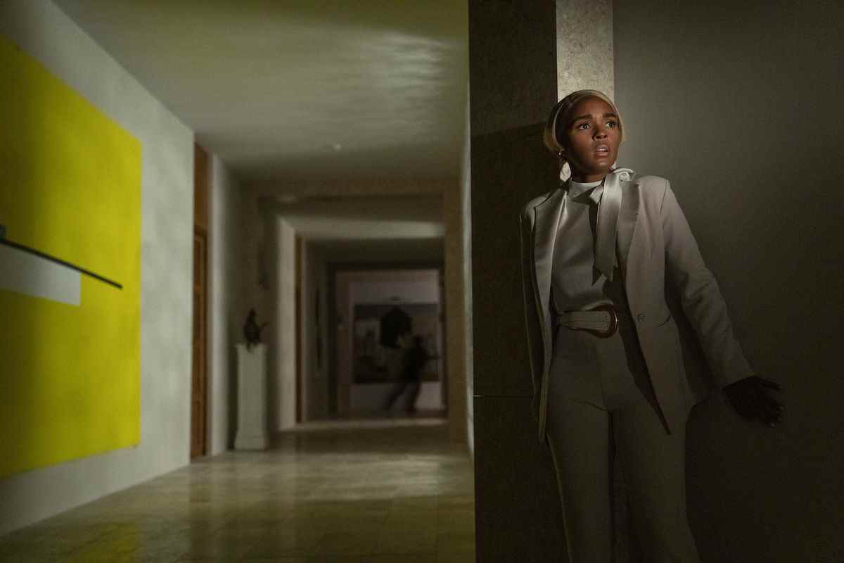 Janelle Monáe a l'air effrayée dans un couloir sombre et vide avec de l'art moderne dans le mur.  En arrière-plan, une silhouette sombre peut juste être vue