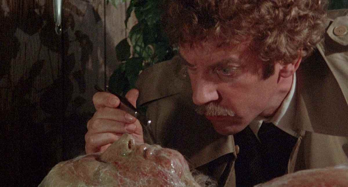 Un homme (Donald Sutherland) examine le visage d'un corps empêtré dans une étrange peau de fibres sinueuses.