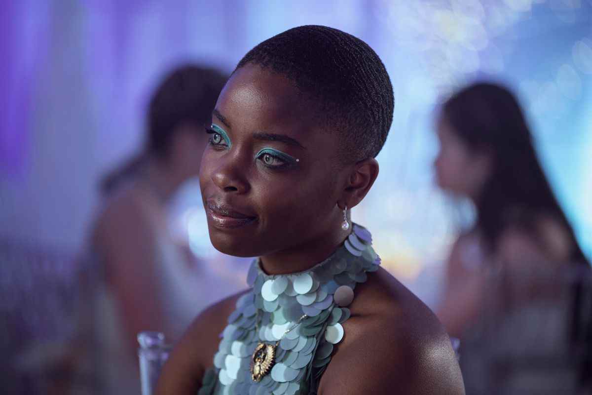 Bianca, une fille noire aux yeux bleu pâle doublés d'un eye-liner bleu argenté assorti, regarde quelqu'un pendant la danse de l'école.  elle porte une tenue bleu argenté scintillante 