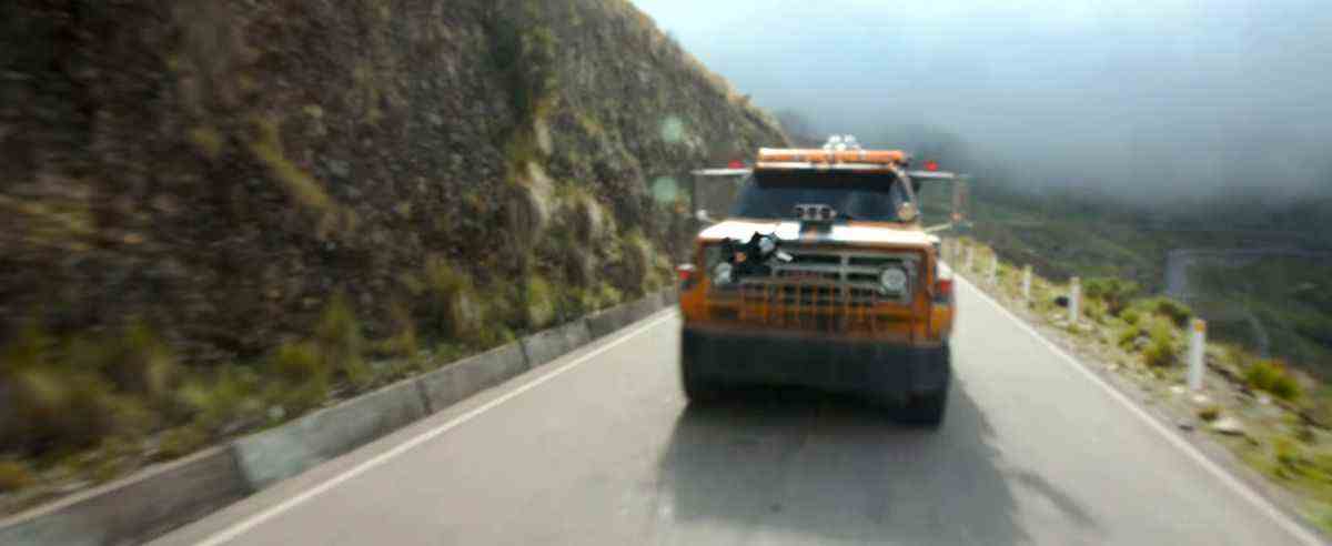 Un transformateur jaune caché sous la forme d'un camion sur une autoroute à flanc de colline tire un missile Transformers: Rise of the Beasts