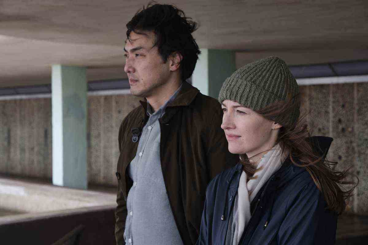 Un homme portant un manteau marron (Takehiro Hira) se tient à côté d'une femme portant un bonnet en tricot gris (Kelly Macdonald) et une veste bleue, ses cheveux étant balayés sur le côté par le vent.