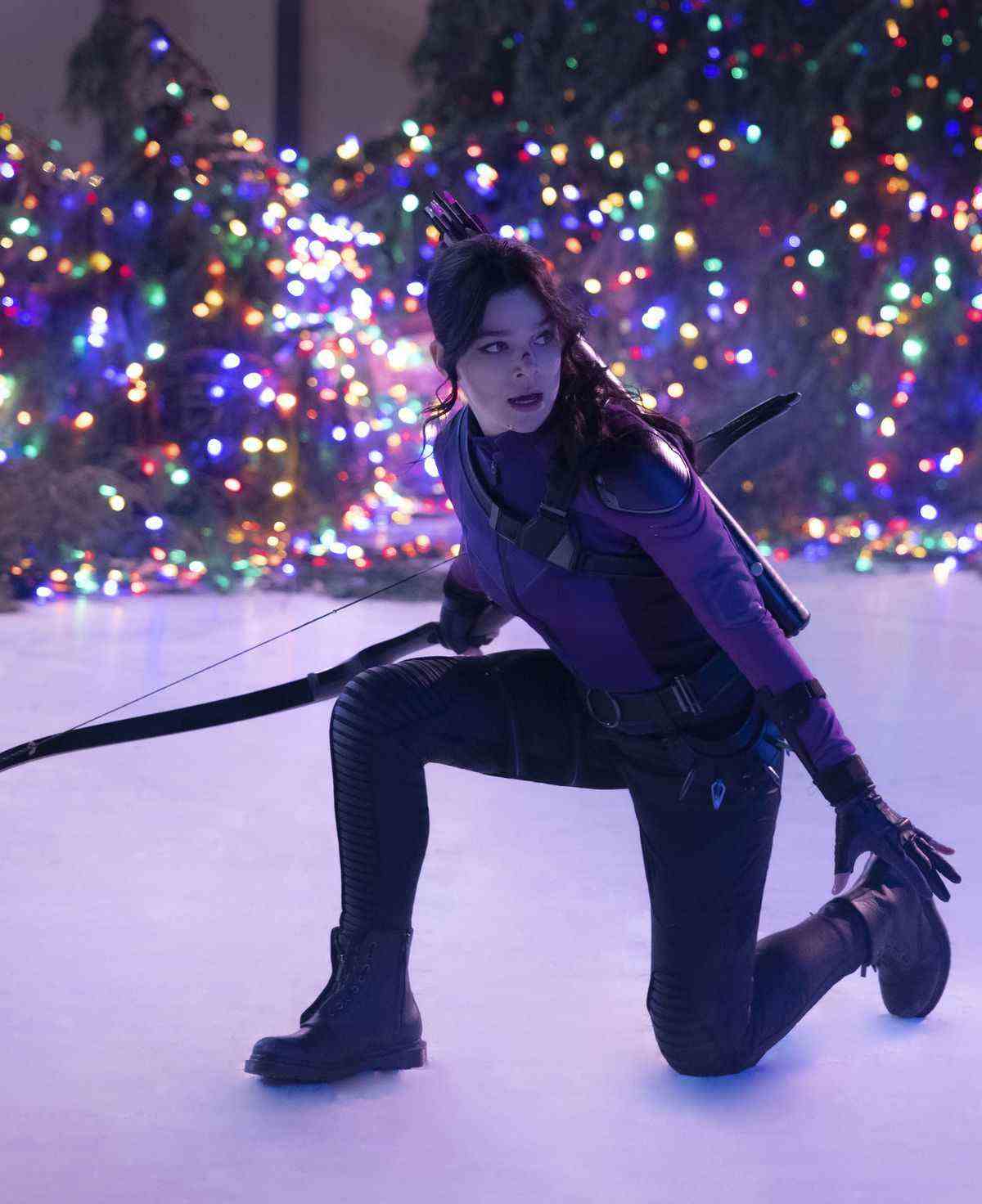 Kate Bishop (Hailee Steinfeld) dans son uniforme violet s'agenouille sur la glace d'une patinoire tout en tenant son arc, tandis que les lumières de Noël l'illuminent par derrière