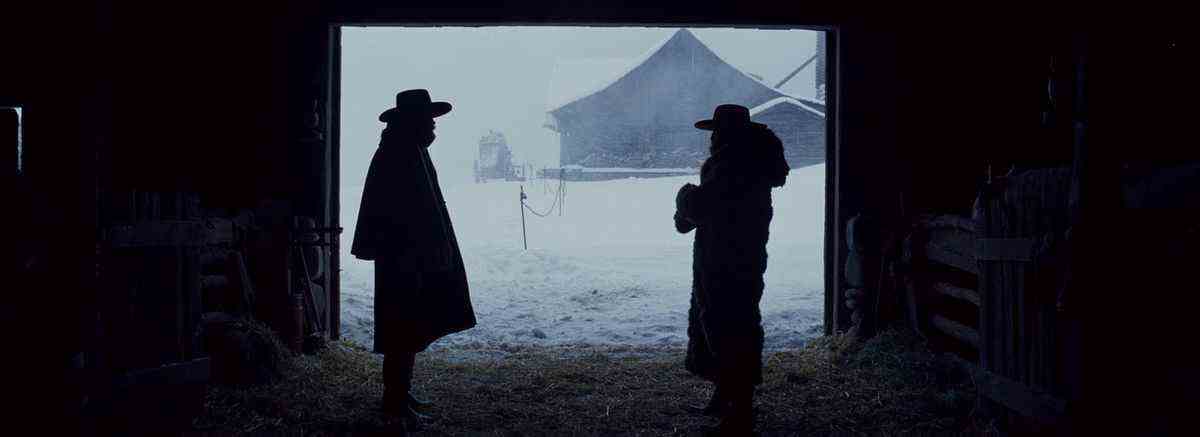 Deux hommes en manteaux d'hiver et chapeaux de cow-boy se tiennent à l'intérieur d'une grange avec un autre bâtiment visible au loin et une tempête de neige à l'extérieur.