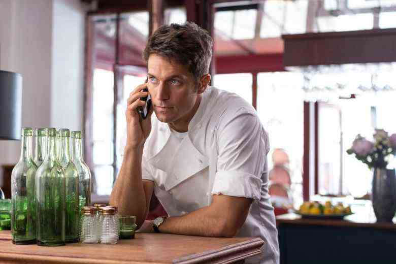 A man in a chef's uniform leaning against a bar, talking on a phone; still from "Emily in Paris"