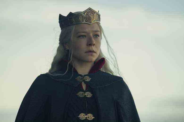 A woman with long, white blond hair, standing outside and wearing a gold crown and dark cloak; still from "House of the Dragon'